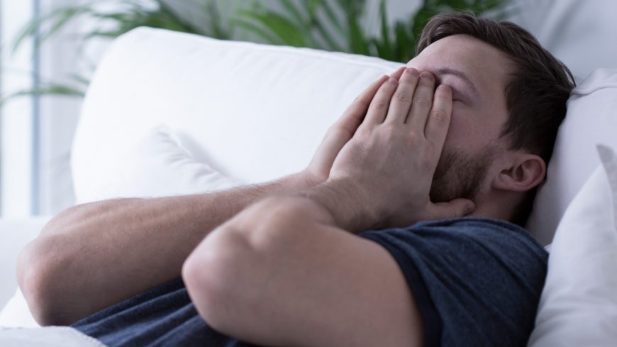 brunette man in navy t-shirt lying on pillow with hands over face